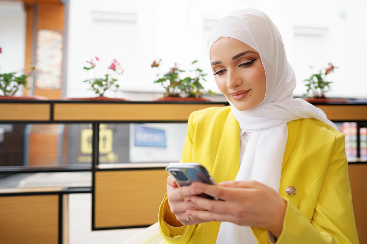 A woman in a white hijab and yellow blazer writing a message on her phone